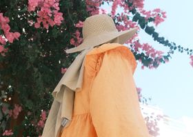 A woman in an orange dress with a sun hat, surrounded by vibrant pink flowers on a sunny day.